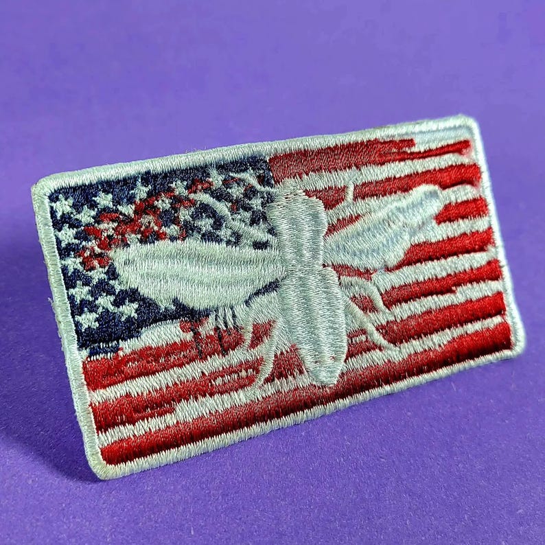 May include: A rectangular embroidered patch featuring a stylized light gray bee centered over a background of the American flag. The flag design includes a blue field with white stars on the left and horizontal red and white stripes on the right.