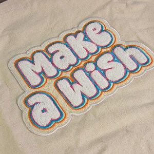 Op de afbeelding: Een cr&egrave;mekleurige canvas draagtas met een grote, getextureerde chenille patch met de woorden "MAKE A WISH" in het wit, omlijnd door lagen felroze, turquoise en oranje draad, wat zorgt voor een opvallend, meerkleurig geborduurd effect.