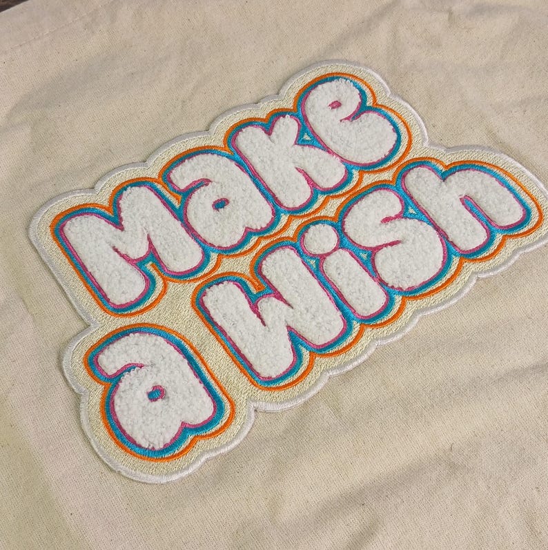 Op de afbeelding: Een cr&egrave;mekleurige canvas draagtas met een grote, getextureerde chenille patch met de woorden "MAKE A WISH" in het wit, omlijnd door lagen felroze, turquoise en oranje draad, wat zorgt voor een opvallend, meerkleurig geborduurd effect.