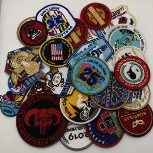 May include: A collection of various embroidered fabric patches in diverse shapes, colors, and sizes. The patches feature assorted graphic designs, including text, logos, symbols, and illustrations, layered together in a dense, overlapping arrangement.