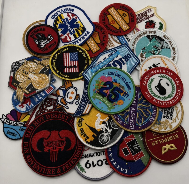May include: A collection of various embroidered fabric patches in diverse shapes, colors, and sizes. The patches feature assorted graphic designs, including text, logos, symbols, and illustrations, layered together in a dense, overlapping arrangement.
