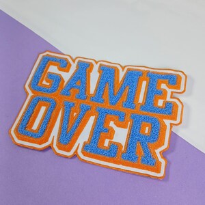 Op de afbeelding: Een rechthoekige geborduurde patch met de tekst GAME OVER in blauwe chenille letters, voorzien van een dikke oranje rand en een witte buitenkant. De patch heeft een getextureerd, pluizig oppervlak en is bedoeld voor op textiel.