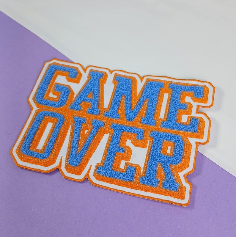 Op de afbeelding: Een rechthoekige geborduurde patch met de tekst GAME OVER in blauwe chenille letters, voorzien van een dikke oranje rand en een witte buitenkant. De patch heeft een getextureerd, pluizig oppervlak en is bedoeld voor op textiel.