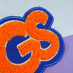 Op de afbeelding: Een chenille patch met de letters GS in feloranje met een witte rand, geplaatst op een donkerblauwe stoffen achtergrond. De letters hebben een getextureerd, pluizig oppervlak en zijn afgewerkt met een dikke blauwe gestikte rand.