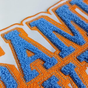 Op de afbeelding: Een chenille patch met de letters A, M en I in een getextureerde lichtblauwe afwerking. De letters zijn omlijnd met een dikke oranje rand op een witte achtergrond, wat zorgt voor een gelaagde uitstraling met zichtbare stiksels.