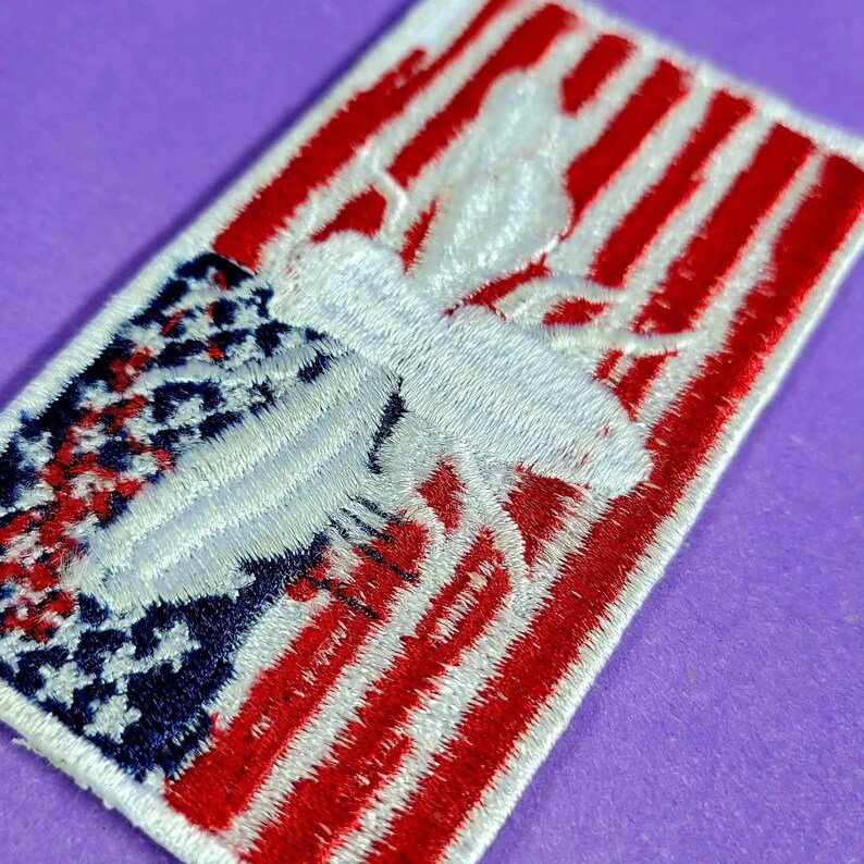May include: A rectangular embroidered patch featuring a white bee silhouette centered over a background of red and white horizontal stripes and a blue corner section with white stars, mimicking the design of the American flag in its entirety.