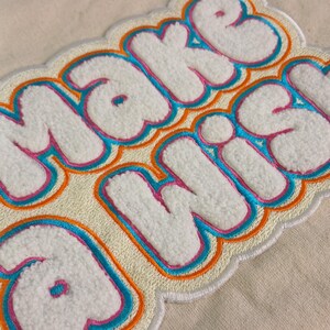 Op de afbeelding: Een witte chenille patch met de tekst "MAKE WISH" in een bubbelachtig lettertype. De letters zijn omlijnd met lagen felroze, turquoise en oranje draad, geplaatst op een cr&egrave;mekleurige stoffen ondergrond met een textuurrijke, pluizige afwerking.