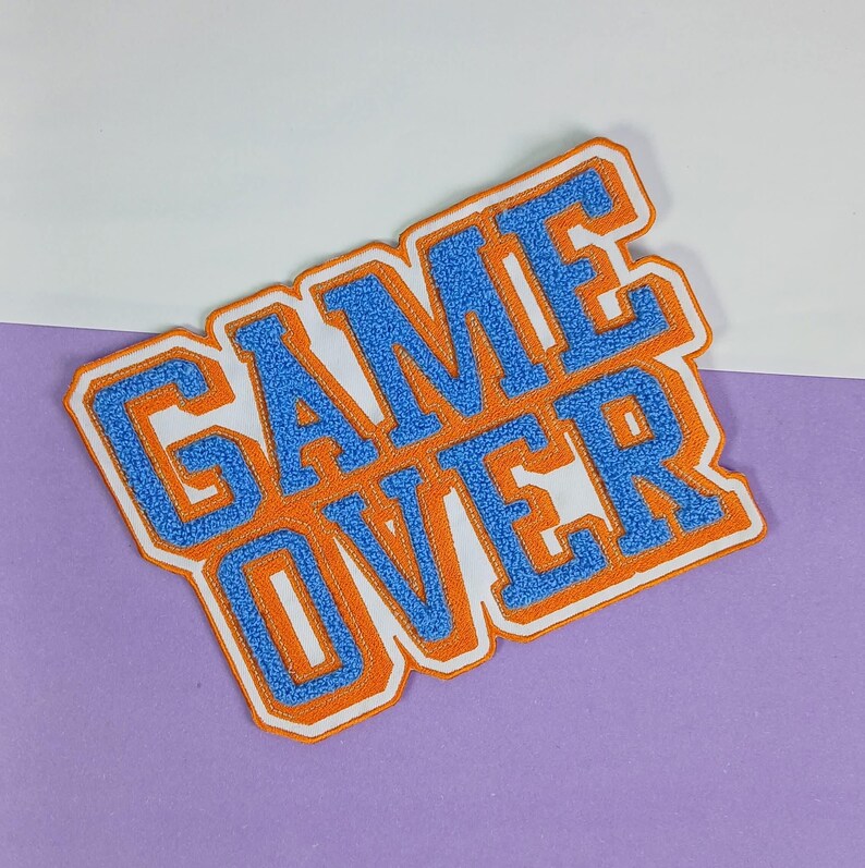 Op de afbeelding: Een stoffen embleem met de tekst GAME OVER in blauwe chenille letters met een dikke oranje rand en een witte omlijning. De letters hebben een getextureerd, pluizig uiterlijk dat kenmerkend is voor borduurwerk in varsity-stijl.