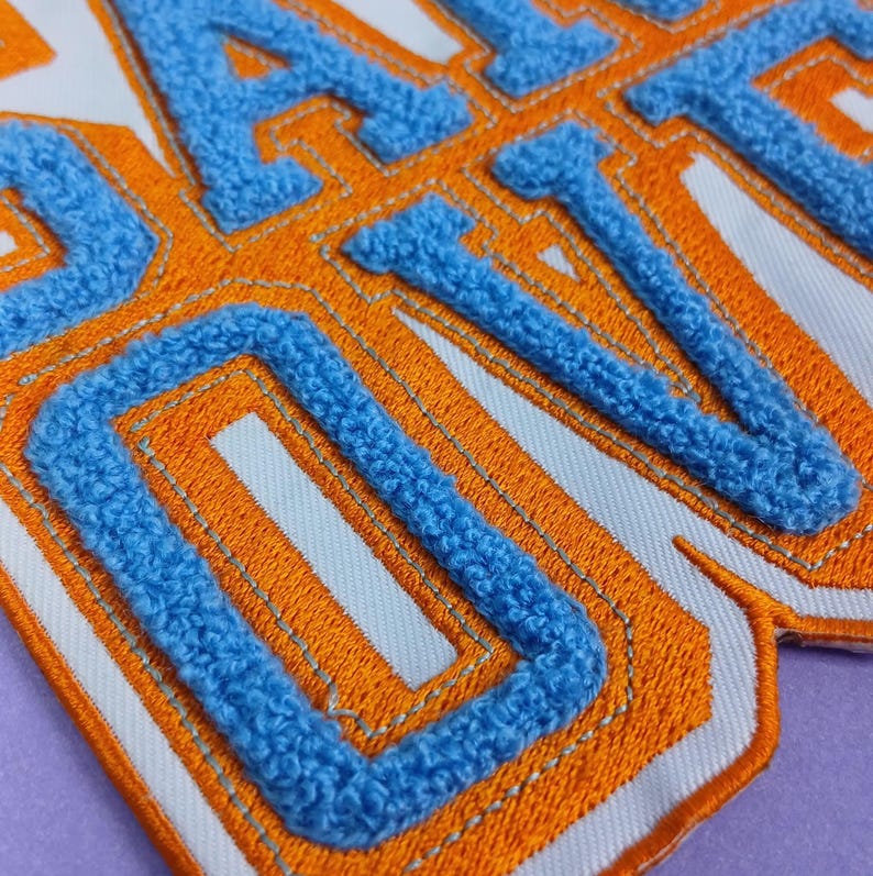 Op de afbeelding: Een chenille patch met de tekst "DAILY DIVA" in lichtblauwe, pluizige, verhoogde letters. De letters zijn omlijnd met een dikke, levendige oranje rand en een dunne zilveren draad, alles op een schone, witte stoffen achtergrond.
