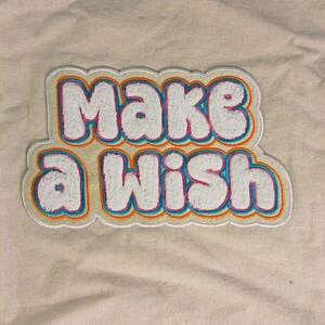 Op de afbeelding: Een chenille patch met de tekst "Make a Wish" in witte, poederige letters. De letters zijn omlijnd met dunne lagen felroze, levendig blauw en oranje, wat een gelaagd, meerkleurig randeffect cre&euml;ert op een beige stoffen achtergrond.