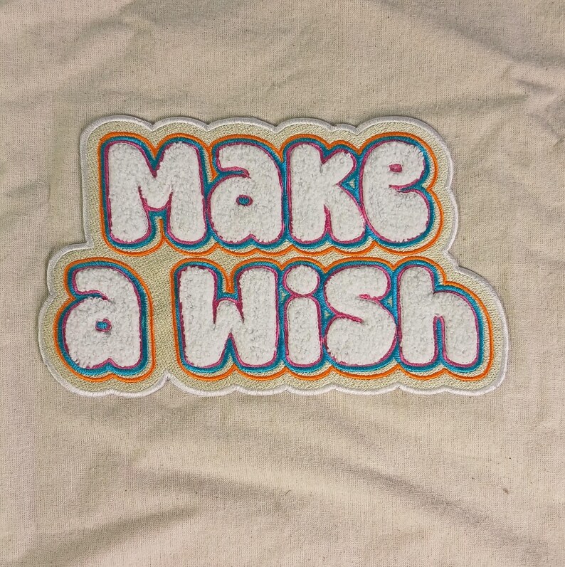 Op de afbeelding: Een chenille patch met de tekst "Make a Wish" in witte, poederige letters. De letters zijn omlijnd met dunne lagen felroze, levendig blauw en oranje, wat een gelaagd, meerkleurig randeffect cre&euml;ert op een beige stoffen achtergrond.