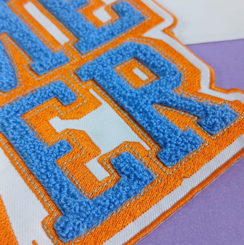 Op de afbeelding: Een chenille patch met de letters "ME" en "ER" in lichtblauw met een dikke oranje rand en een witte achtergrond. De getextureerde, pluizige letters zijn omlijnd met zilverkleurig stiksel, wat zorgt voor een gelaagde, varsity-stijl uitstraling.