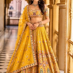 May include: A vibrant yellow lehenga choli set featuring intricate multi-colored embroidery, small mirror work accents, and a matching sheer dupatta. The ensemble includes a cropped blouse with short sleeves and a voluminous, floor-length skirt.