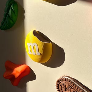 Könnte beinhalten: Eine Sammlung von Wanddekorationsobjekten, bestehend aus einem gelben kreisförmigen Teil mit einem weißen Buchstaben m, einem grünen blattförmigen Element, einer orangefarbenen abstrakten Form und einem braunen ovalen Keks mit floralen Mustern.