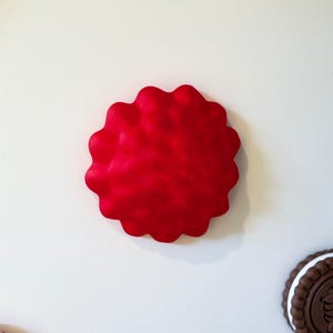 Puede incluir: Un objeto circular de color rojo brillante con textura y borde festoneado. La superficie presenta una serie de protuberancias redondeadas en relieve. Una parte de una galleta tipo sándwich marrón y una galleta con glaseado rosa son visibles.