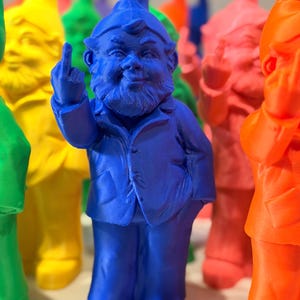 May include: A bright blue 3D printed gnome figurine wearing a jacket and hat, standing with one hand raised in a middle finger gesture. The gnome has a detailed beard and facial features, surrounded by other colourful gnomes in the background.