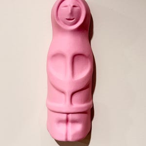 May include: A solid pink, matte-finished sculpture of a stylised human figure. The figure features a rounded head with a simple carved face, a hooded garment, and a smooth, elongated body with subtle indentations representing arms and legs.