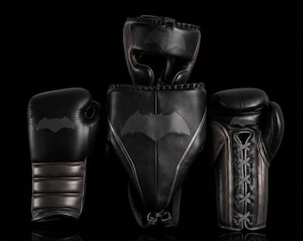 Könnte beinhalten: Ein Set schwarzer Boxausrüstung bestehend aus einem Kopfschutz, einem Tiefschutz und zwei Boxhandschuhen. Jedes Teil ist aus schwarzem Leder gefertigt und zeigt ein dunkelgraues Batman-Logo, das mittig auf der Vorderseite angebracht ist.