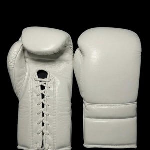 May include: A pair of white boxing gloves. The left glove features a lace-up closure system with white laces running through reinforced eyelets. The right glove shows the smooth, solid white exterior of the back of the hand and wrist area.