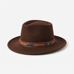 Könnte beinhalten: Ein dunkelbrauner Filzhut im Fedora-Stil mit strukturierter Krone und breiter Krempe. Der Hut ist mit einem dunkelbraunen Lederband verziert, das eine dekorative orangefarbene Lederschnürung und einen kleinen Metallniet an der Seite aufweist.