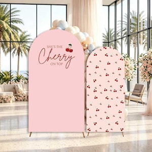 May include: Two arched event backdrops in light pink. The larger left panel features the text "SHE'S THE Cherry ON TOP" in dark red script and print with a small cherry graphic. The smaller right panel has a repeating pattern of small red cherries.