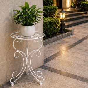 Op de afbeelding: Een witte metalen plantenstandaard met een rond blad en gebogen, krulvormige poten. De standaard bevat een ronde, glanzend witte pot met een groene plant met puntige bladeren, geplaatst op een betegeld pad van natuursteen.