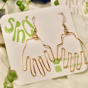 May include: A pair of gold-toned wire earrings shaped like open hands, suspended from standard earring hooks. The earrings are attached to a white display card featuring the green text "shop spinc" and social media icons.