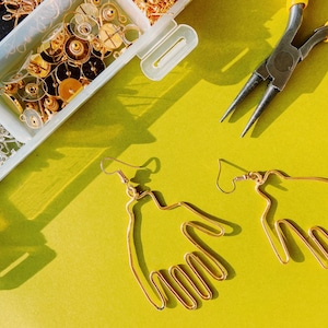May include: Two gold-toned earrings shaped like hands with five fingers. The earrings are on a yellow surface.
