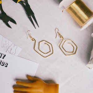 Op de afbeelding: Een platte lay-out van sieradenmaakbenodigdheden, waaronder twee gouden draad oorbellen, twee spoelen draad en twee tangen. De afbeelding bevat ook een stuk papier met de tekst "Tiny Hands Art" erop gedrukt.