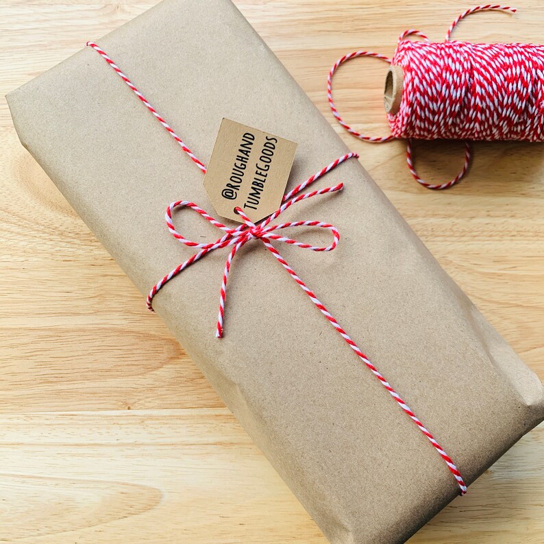 May include: A brown paper package tied with red and white baker's twine. A small brown tag is attached to the twine with the text "@ROUGHANDTUMBLEGOODS".