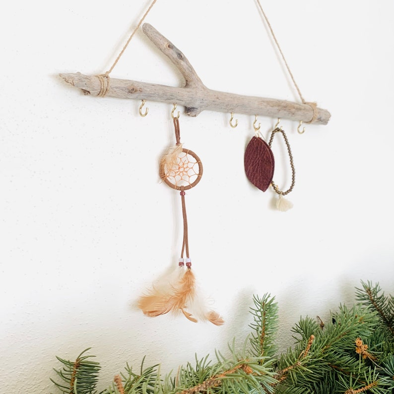 May include: A driftwood branch with hooks for hanging jewelry, featuring a dreamcatcher with brown leather and feathers, a brown leather leaf, and a brown beaded bracelet.