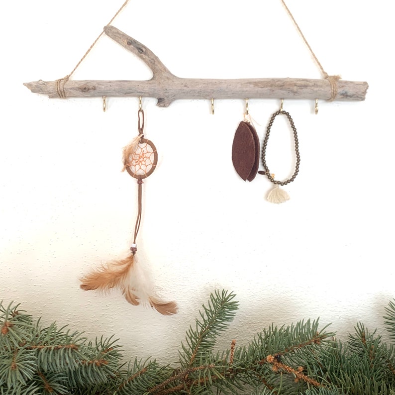 May include: A rustic wooden branch with five hooks, used as a jewelry organizer. A dreamcatcher, a brown felt leaf, and a beaded necklace hang from the hooks.