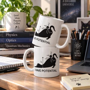 Peut inclure: Deux tasses en céramique blanche empilées verticalement, chacune présentant un graphique noir d'un chat dans une pose d'étirement avec des diagrammes de force physique et le texte "I HAVE POTENTIAL" imprimé en noir sur la surface.