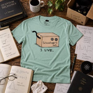 May include: A light green t-shirt featuring a graphic of a cardboard box labeled "Schrödinger's cat" with a cat tail sticking out and the text "I LIVE." printed below the box. The design is centered on the chest of the short-sleeved garment.