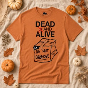 May include: A burnt orange short-sleeve t-shirt featuring a black graphic of a cardboard box with a cat peeking out. The text reads DEAD X AND ALIVE above the box and DO NOT OBSERVE on the box, with the words SCHRODINGER CAT written on the side.