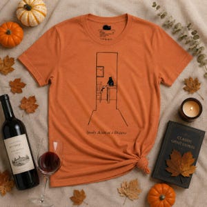 Puede incluir: Una camiseta de manga corta color naranja quemado con un dibujo lineal negro de un gato sentado sobre una mesa dentro de un marco de puerta. Debajo de la ilustración, el texto dice Spooky Action at a Distance. Tiene cuello redondo.