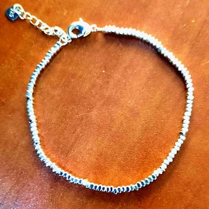 May include: A delicate silver-toned beaded bracelet featuring small, textured metallic beads strung along a thin wire. The bracelet includes a lobster claw clasp, a short extender chain, and a small rectangular tag stamped with the number 925.