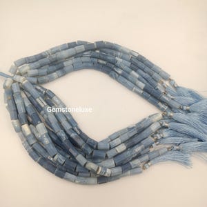 May include: A collection of multiple strands of cylindrical blue gemstone beads, each featuring a smooth, polished finish with varying shades of light and dark blue. The strands are finished with light blue fabric tassels and the text Gemstoneluxe.