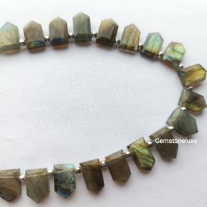 Op de afbeelding: Een streng gefacetteerde labradoriet kralen in de vorm van vijfhoekige schilden, elk met iriserende flitsen van blauw, groen en goud. Kleine metalen tussenkralen zijn tussen elke steen geplaatst. De tekst Gemstoneluxe is zichtbaar.