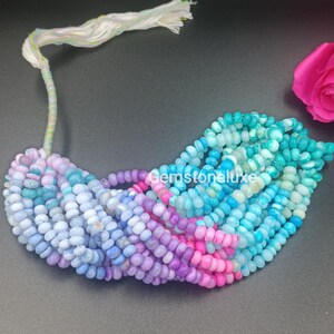 Puede incluir: Una hebra de pequeñas cuentas redondas en forma de disco en un degradado de colores que va desde el morado claro y azul hasta el turquesa brillante y verde. Las cuentas están ensartadas en un cordón blanco con una borla. El texto Gemstoneluxe es visible.
