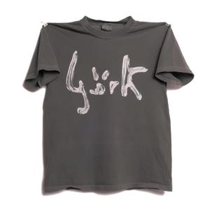 May include: A charcoal gray short-sleeve t-shirt featuring a white, brushstroke-style graphic print on the chest that reads "Björk". The garment has a crew neckline and a straight hem, with small metal clips visible at the top shoulder seams.