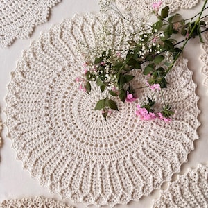 May include: A collection of round, off-white crocheted doilies featuring intricate, radiating patterns with scalloped edges. A sprig of small pink flowers and delicate white baby's breath is placed across the center of the primary doily.