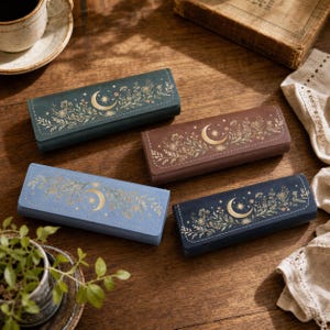 Op de afbeelding: Vier rechthoekige etuis in donkergroen, bruin, lichtblauw en marineblauw. Elk etui is voorzien van een goudkleurig motief van een halve maan en ster, omringd door delicate botanische illustraties van bladeren en kleine bloemen op de klep.