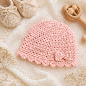 Könnte beinhalten: Eine hellrosa gehäkelte Babymütze mit strukturiertem Stichmuster, gewelltem Rand und einer kleinen gehäkelten Schleife an der Seite. Die Mütze wird von einem Paar passender beiger gehäkelter Babyschuhe und einer Holzrassel begleitet.