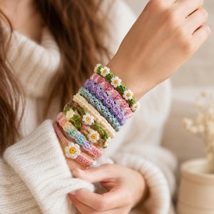 May include: A stack of eight handmade crochet bracelets worn on a wrist. Each bracelet features a unique color scheme, including shades of green, pink, purple, blue, and cream, with small white and yellow flower motifs integrated into the woven design.
