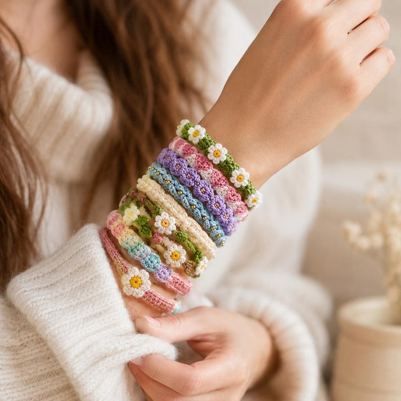 May include: A stack of eight handmade crochet bracelets worn on a wrist. Each bracelet features a unique color scheme, including shades of green, pink, purple, blue, and cream, with small white and yellow flower motifs integrated into the woven design.