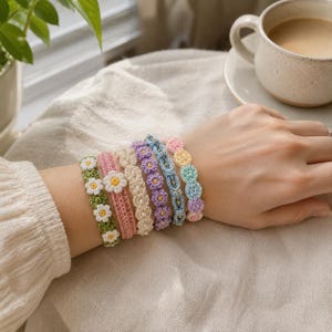 May include: A collection of six handmade crochet bracelets featuring various designs, including white daisy flowers on green, a pink band with a white flower, cream beads, purple flowers, blue with gold beads, and a pastel multi-colored scalloped pattern.