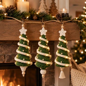 Op de afbeelding: Drie kegelvormige kerstboomornamenten van groen haakgaren, elk bekroond met een witte gehaakte ster en versierd met een spiraalvormige witte haakguirlande, kleine witte kralen en geïntegreerde warmwitte kerstverlichting.