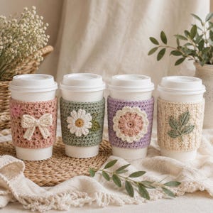 May include: Four white disposable coffee cups with crochet sleeves. The first sleeve is dusty pink with a cream bow. The second is sage green with a white daisy. The third is lavender with a pink and cream flower. The fourth is cream with green leaves.
