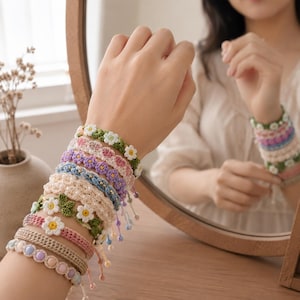 May include: A stack of multiple handmade crochet bracelets featuring various designs, including white daisy flowers with yellow centers, green leaves, and pastel-colored beads. The bracelets are crafted with intricate yarn patterns in shades of green, pink, purple, blue, and cream.